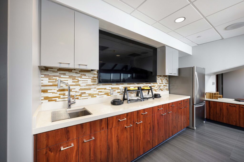A kitchen kitchenette with wood-grain lower cabinets, white countertops, a sink, a wall-mounted television, and a stainless steel refrigerator.