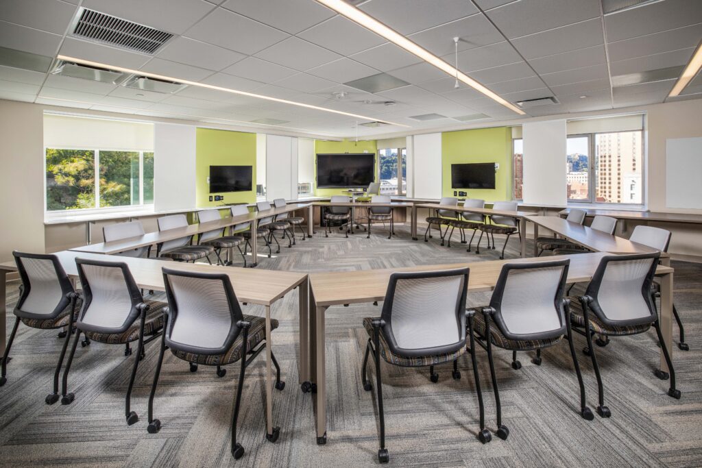 A conference room with a U-shaped arrangement of desks and chairs facing three wall-mounted television screens.