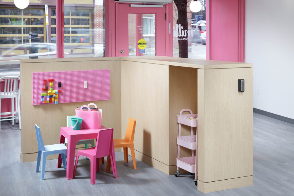 A children’s play area with a small pink table, chairs in blue, pink, and orange, a wall-mounted pink pegboard with building blocks, and a rolling metal cart.