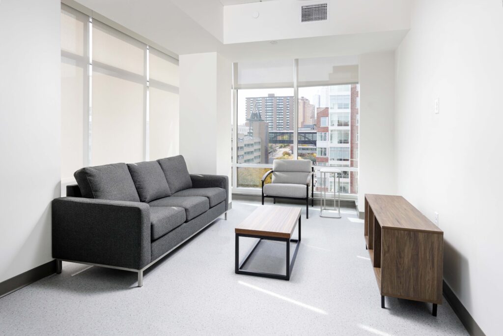 A furnished living room with a grey sofa, a matching armchair, a wooden coffee table, and a media console, featuring large windows looking out onto a city skyline.