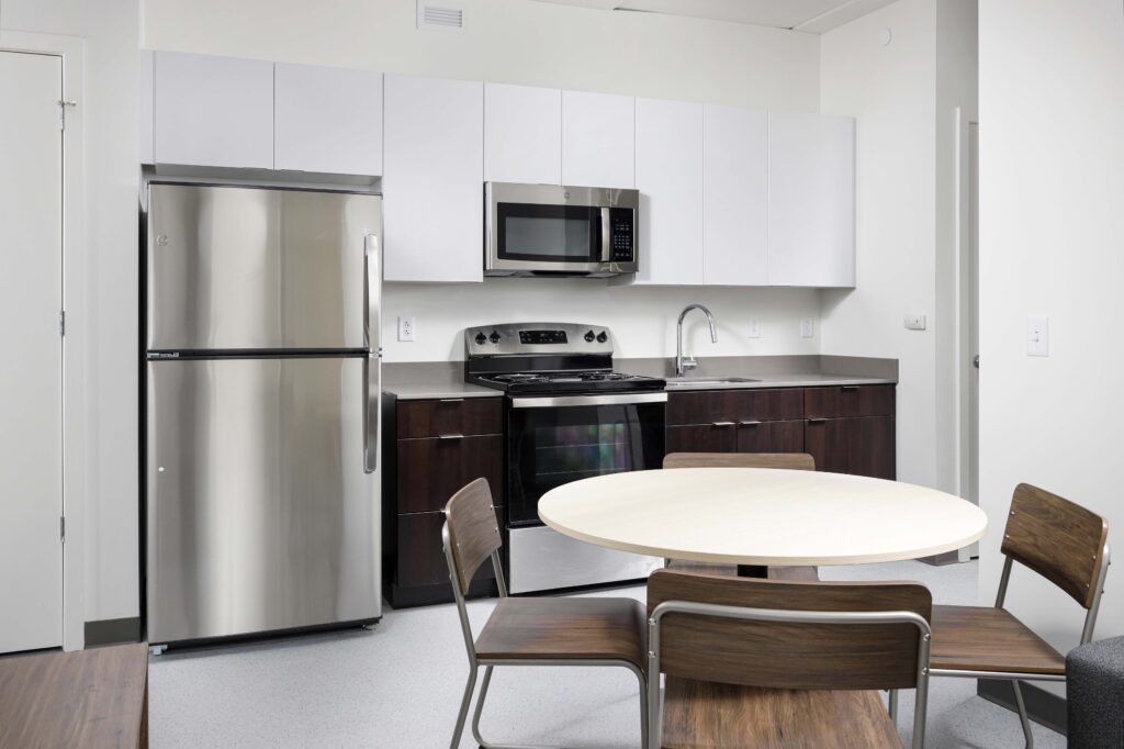 A kitchenette with white upper cabinets, dark wood lower cabinets, a stainless steel refrigerator and stove, and a round wooden dining table with four chairs.