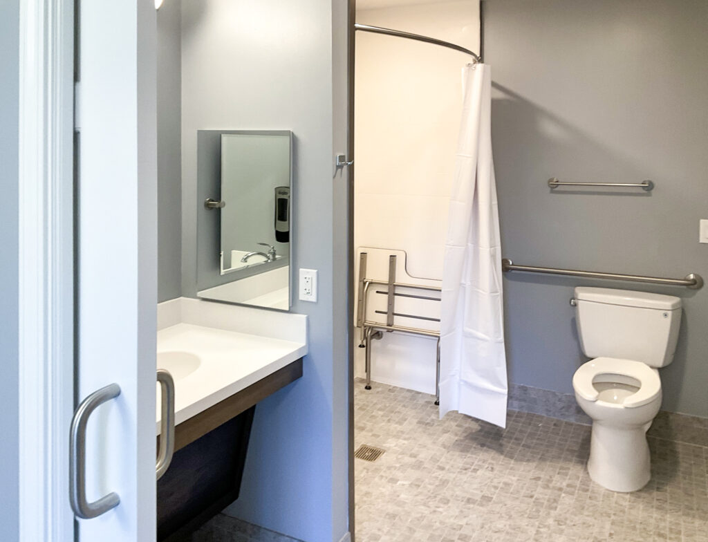 Accessible bathroom with a wall-mounted sink, a roll-in shower with a fold-down seat and curtain, and a toilet with grab bars.