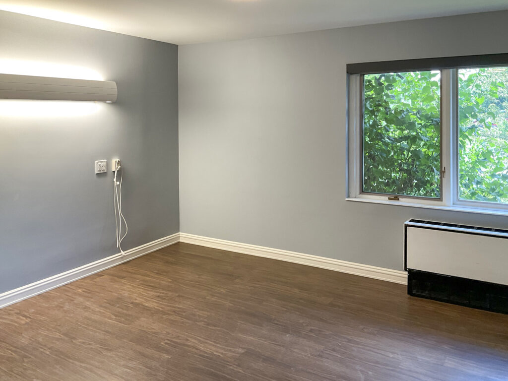 An empty room with light gray walls, wood-look flooring, a window overlooking greenery, a wall-mounted light fixture, and an under-window HVAC unit.
