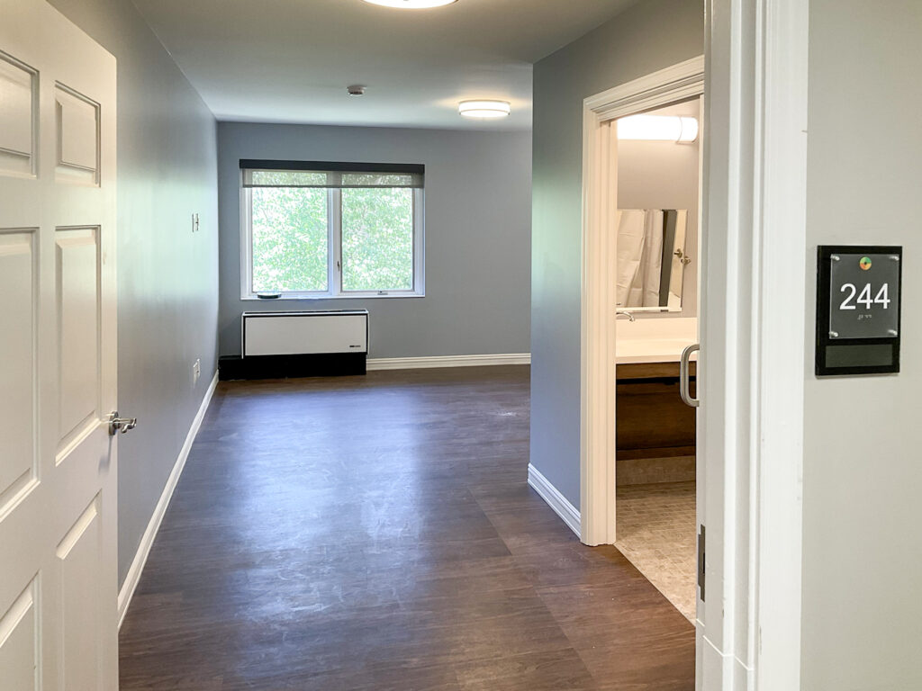 Interior view of a hotel room featuring a bedroom area with a window, a bathroom entrance to the right, and a sign labeled “244” on the wall.