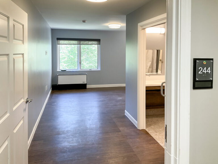 Interior view of a hotel room featuring a bedroom area with a window, a bathroom entrance to the right, and a sign labeled “244” on the wall.