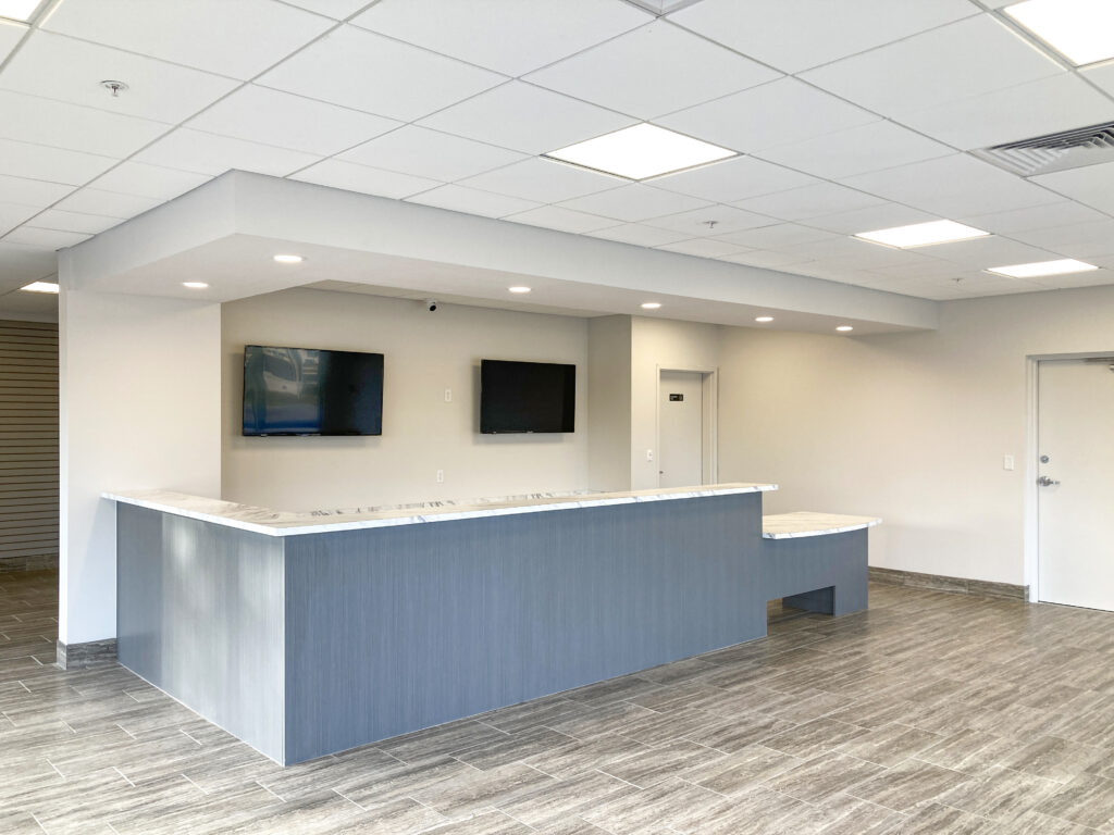 A modern blue reception desk with a white countertop and two wall-mounted televisions in a bright office lobby.