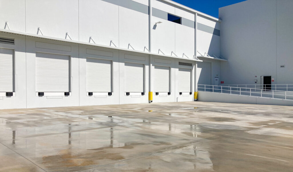 A row of closed loading dock doors on a white industrial building with a wet concrete apron.
