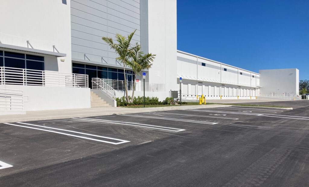 A modern white warehouse building with a large parking lot, accessible entrance, and palm tree.