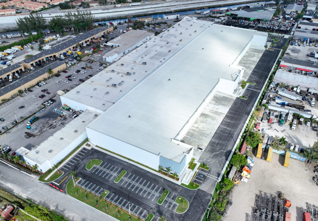 Aerial view of a large white industrial warehouse with a parking lot and surrounding shipping docks.