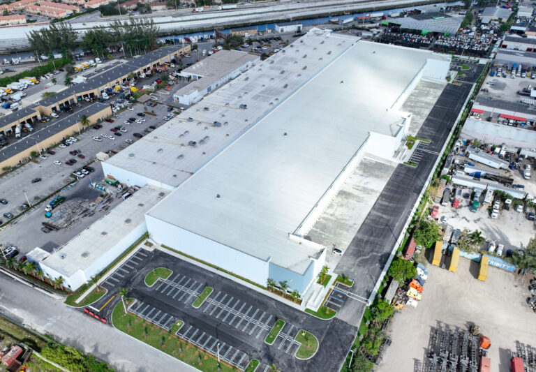 Aerial view of a large white industrial warehouse with a parking lot and surrounding shipping docks.