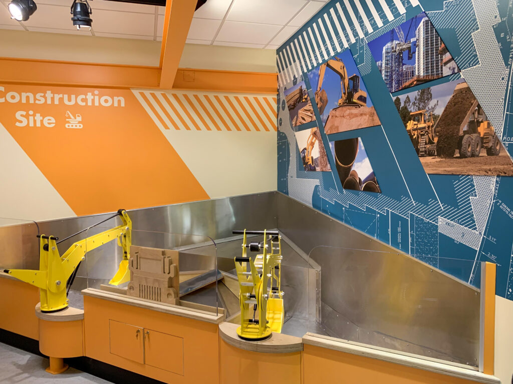 Two yellow construction-themed play excavators installed at a counter in a children’s museum exhibit titled “Construction Site.”