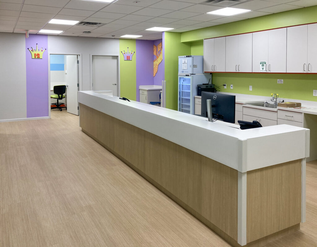 A pediatric reception desk with a white counter, light wood front, and cabinets, featuring colorful walls with crown-shaped room numbers.