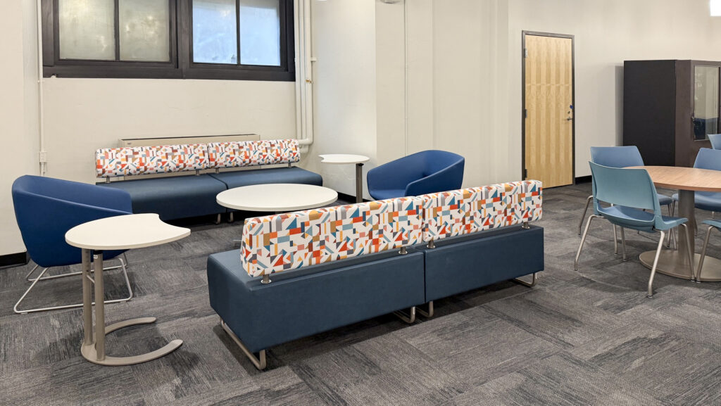 A lounge area with two blue sofas featuring patterned back cushions, blue armchairs, and several small tables on a grey carpeted floor.