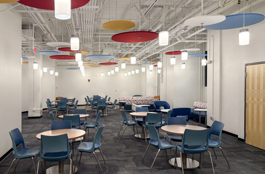 A modern cafeteria or breakout space with several circular tables, blue chairs, and colorful acoustic ceiling panels hanging above.