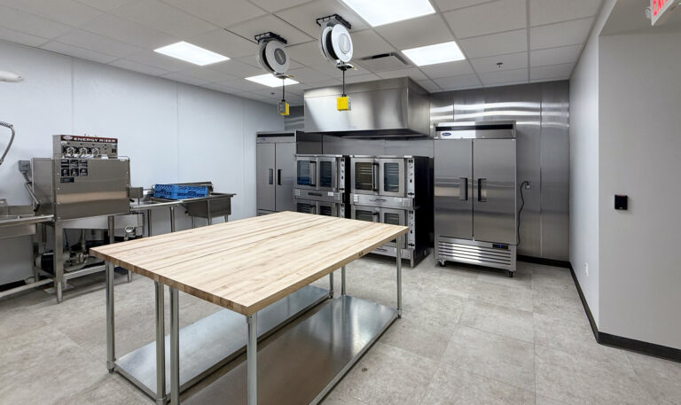 Commercial kitchen with a wooden prep table, stainless steel refrigerators, stacked ovens, and an industrial mixer.