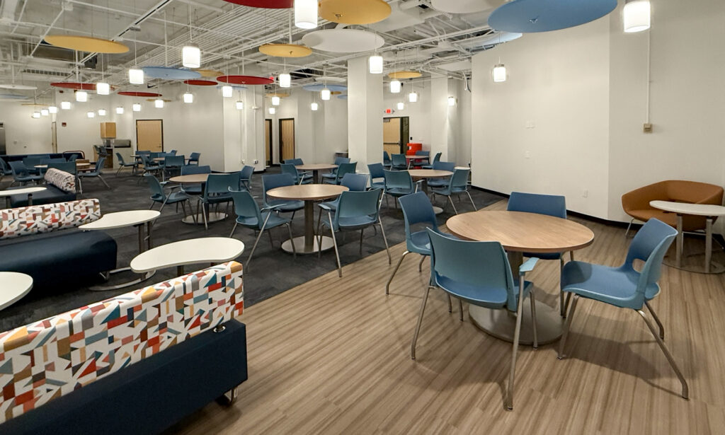 A large open breakroom with circular tables, blue chairs, and patterned sofas beneath colorful circular ceiling acoustic panels.