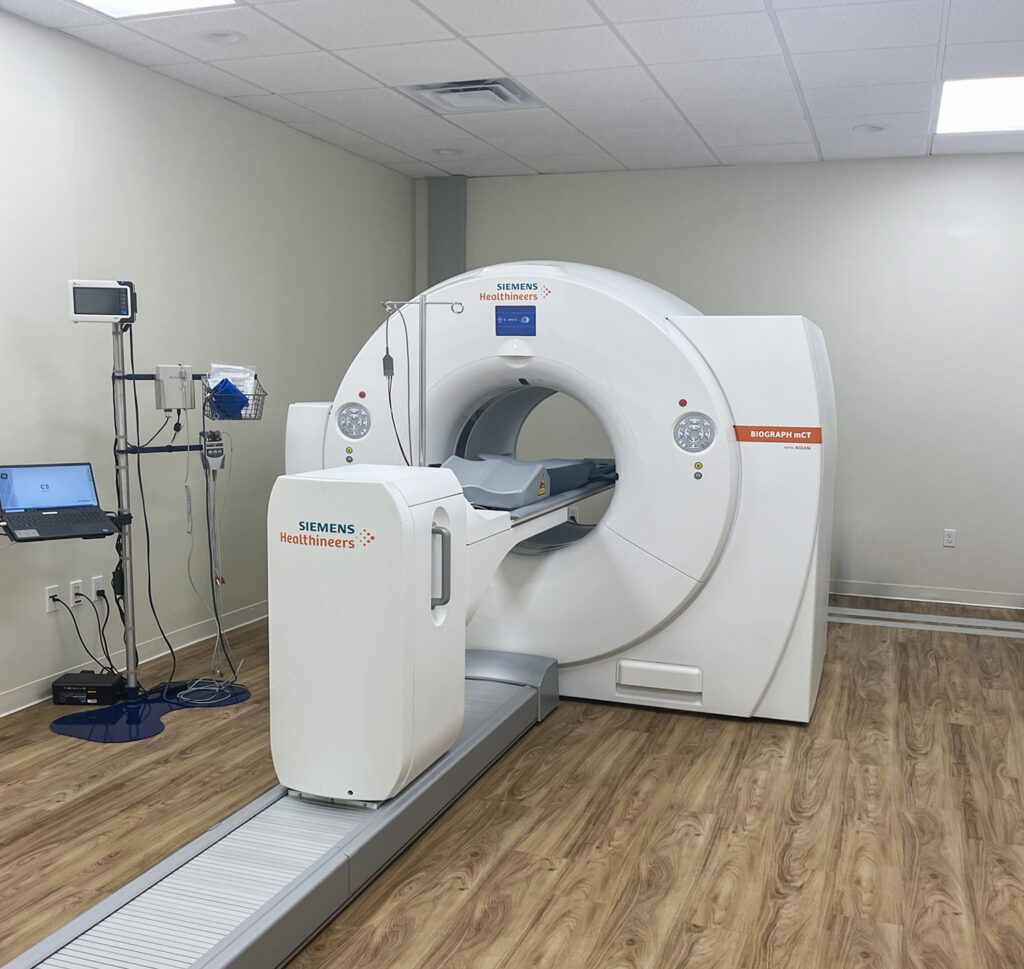 A Siemens Healthineers Biograph mCT PET/CT scanner in a clinical room with a laptop on a cart.