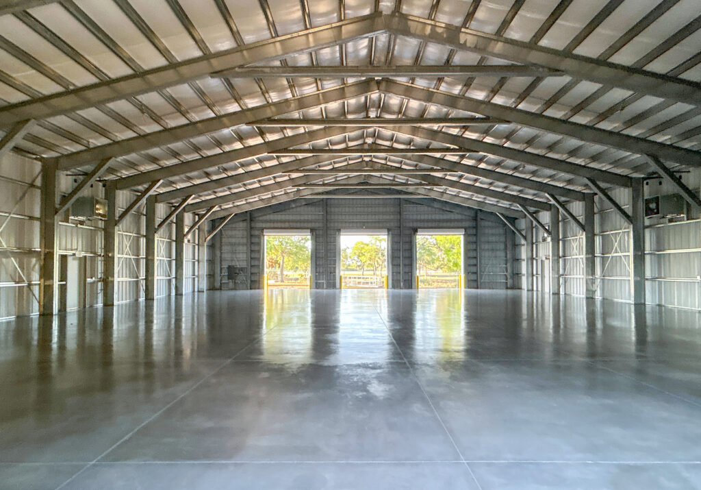 The interior of a large, empty metal warehouse with a polished concrete floor and three open doorways looking out onto trees.
