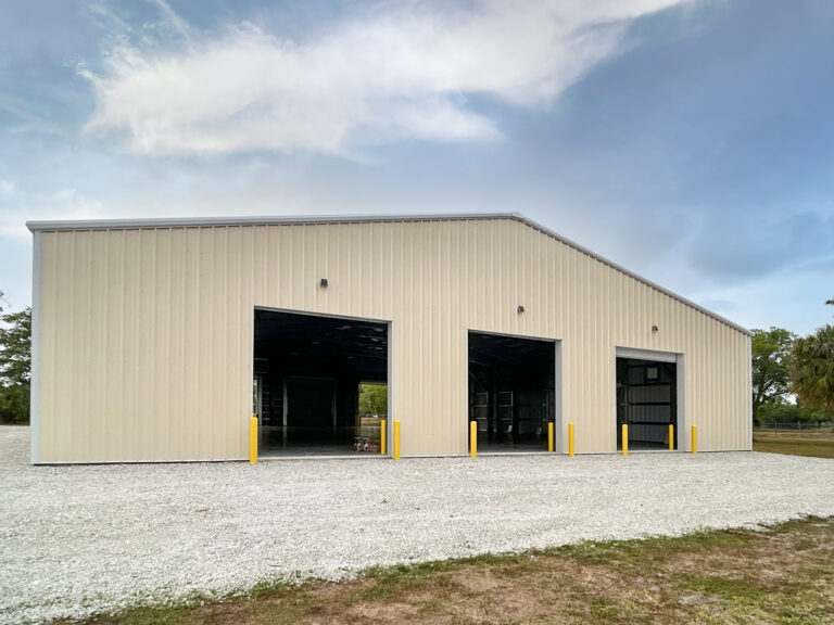 A cream-colored metal building with three open garage bays and yellow safety bollards, situated on a gravel lot.