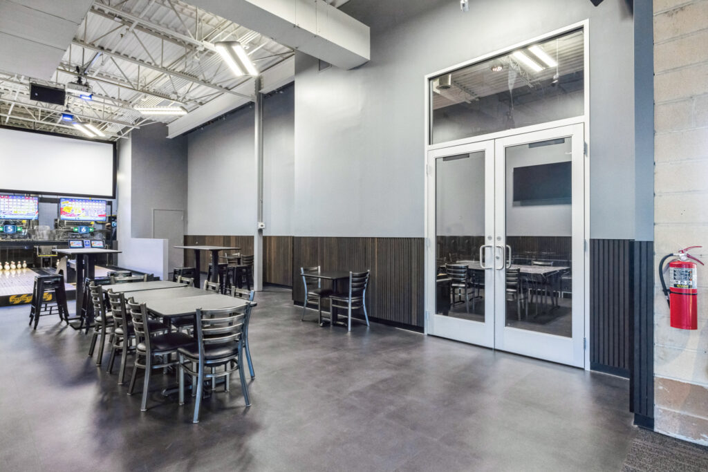 Bowling alley interior featuring dining tables and chairs, a glass-enclosed room, and a fire extinguisher on the wall.