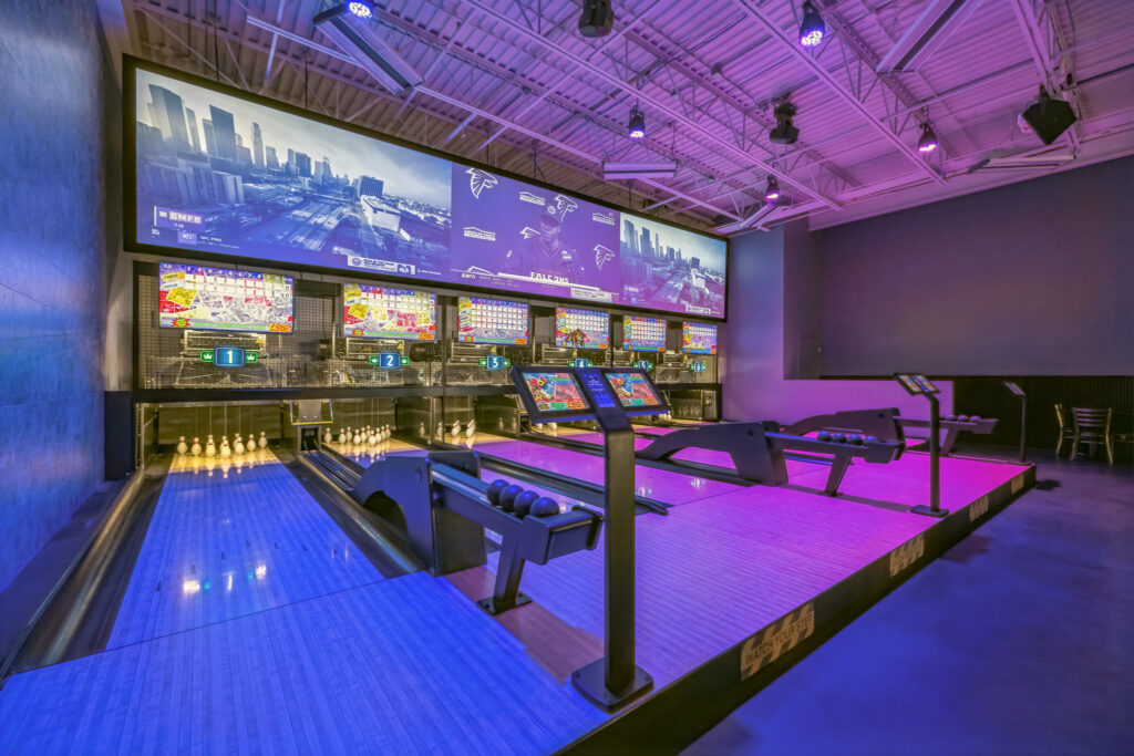 A modern bowling alley with multiple lanes, scoring screens, and a large projection screen displaying sports broadcasts under purple lighting.