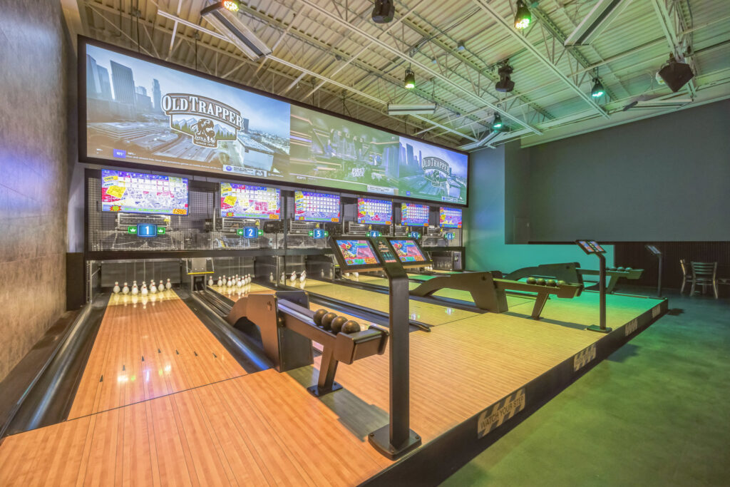 Bowling alley featuring multiple lanes with ball returns and digital scoreboards beneath large overhead screens.