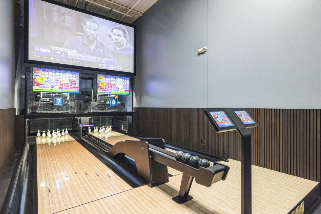Two bowling lanes with pins and ball returns, featuring scoring screens and a large television mounted above.