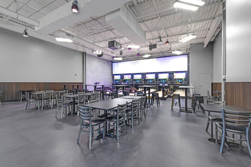 Bowling alley with a lounge area featuring multiple tables and chairs facing the lanes and digital scoring screens.