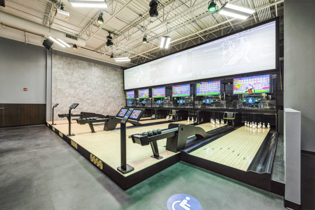 An indoor bowling alley with multiple lanes, each featuring an individual touchscreen console and a large overhead screen displaying sports.