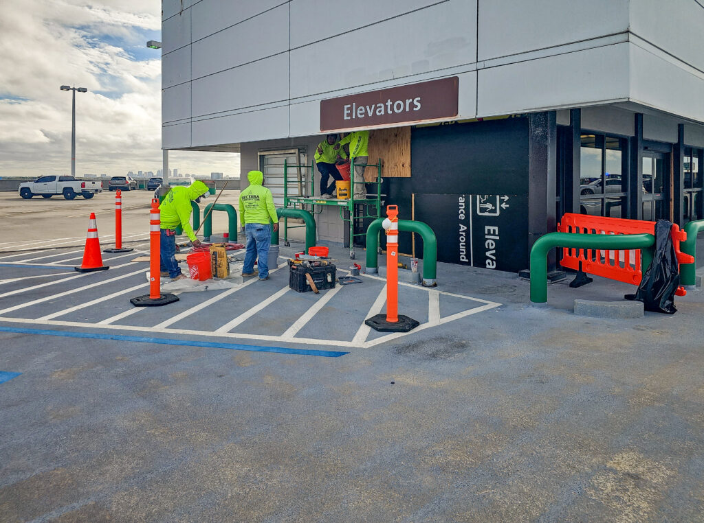Construction workers in neon yellow safety vests perform repairs at an elevator entrance in a parking garage.