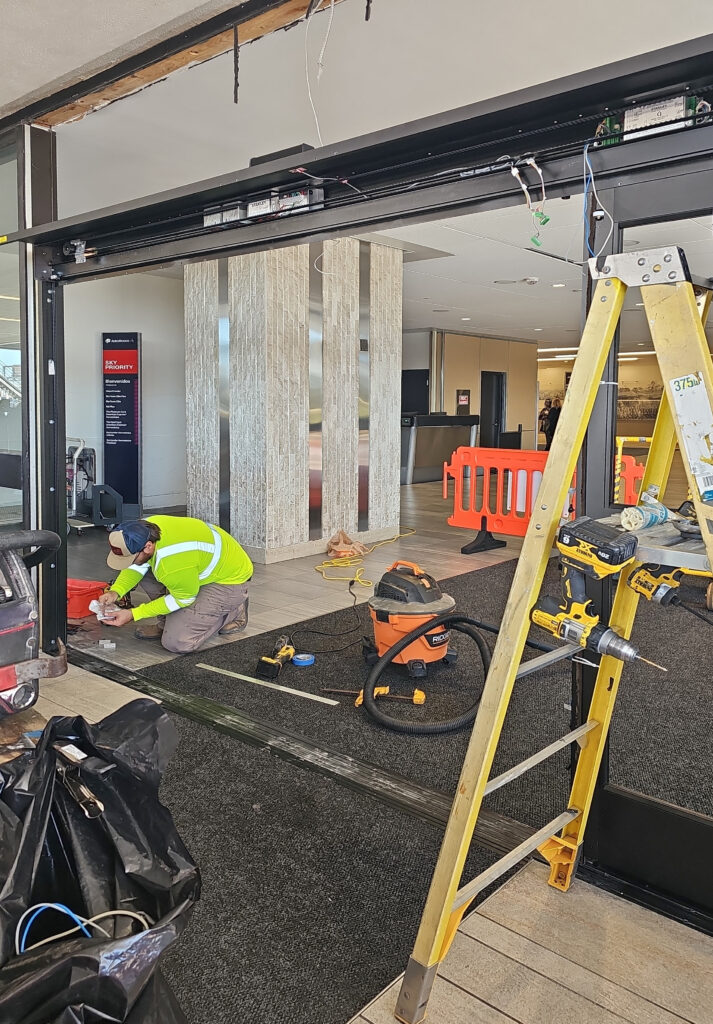 A technician in a high-visibility vest repairs the track of a sliding glass door at an airport entrance, with a ladder and tools nearby.