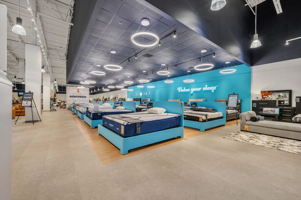 A mattress showroom with rows of beds on blue frames, bright teal walls, and circular pendant lights.
