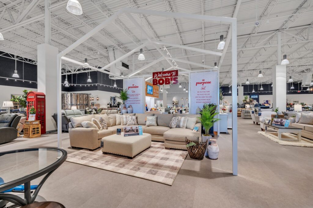 A sectional sofa and ottoman arranged on a plaid rug inside a furniture store, framed by a white house-shaped display structure.