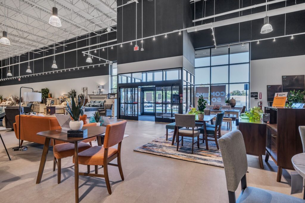A furniture store showroom featuring dining sets, lounge chairs, and sofas in an open, brightly lit space with large glass doors at the entrance.