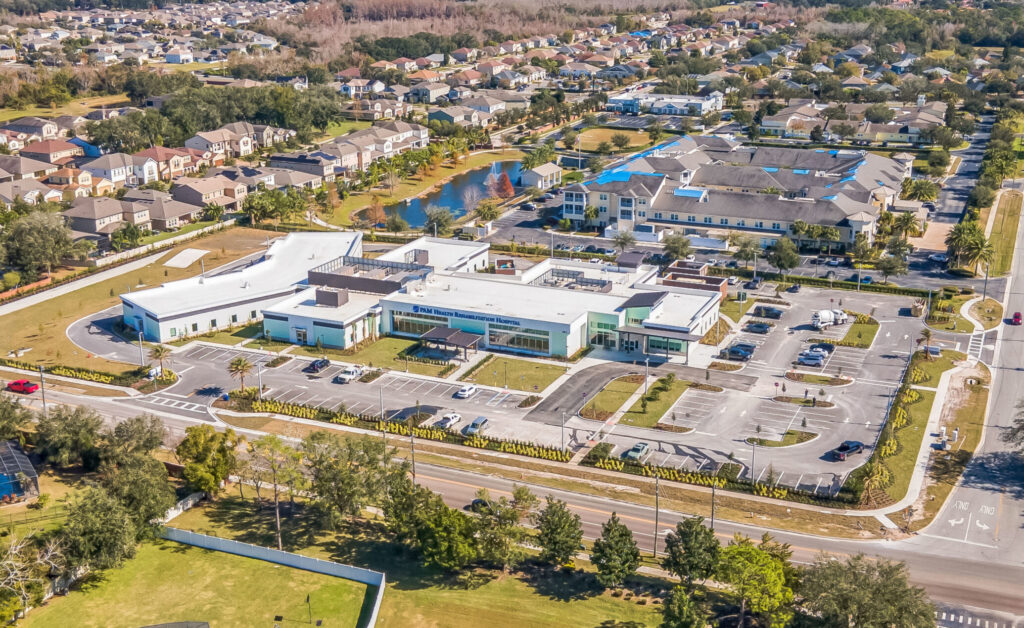 Aerial view of the PAM Health Rehabilitation Hospital building, its parking lot, and the surrounding suburban neighborhood.