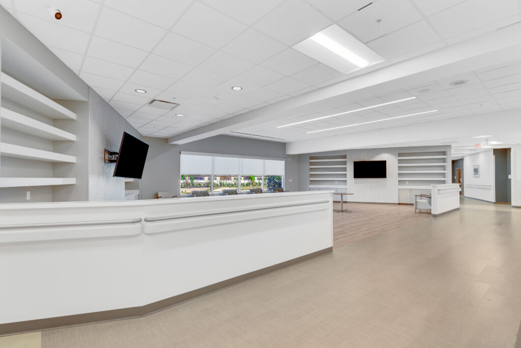 A modern reception area with a large white curved desk, light wood flooring, and wall-mounted television screens.
