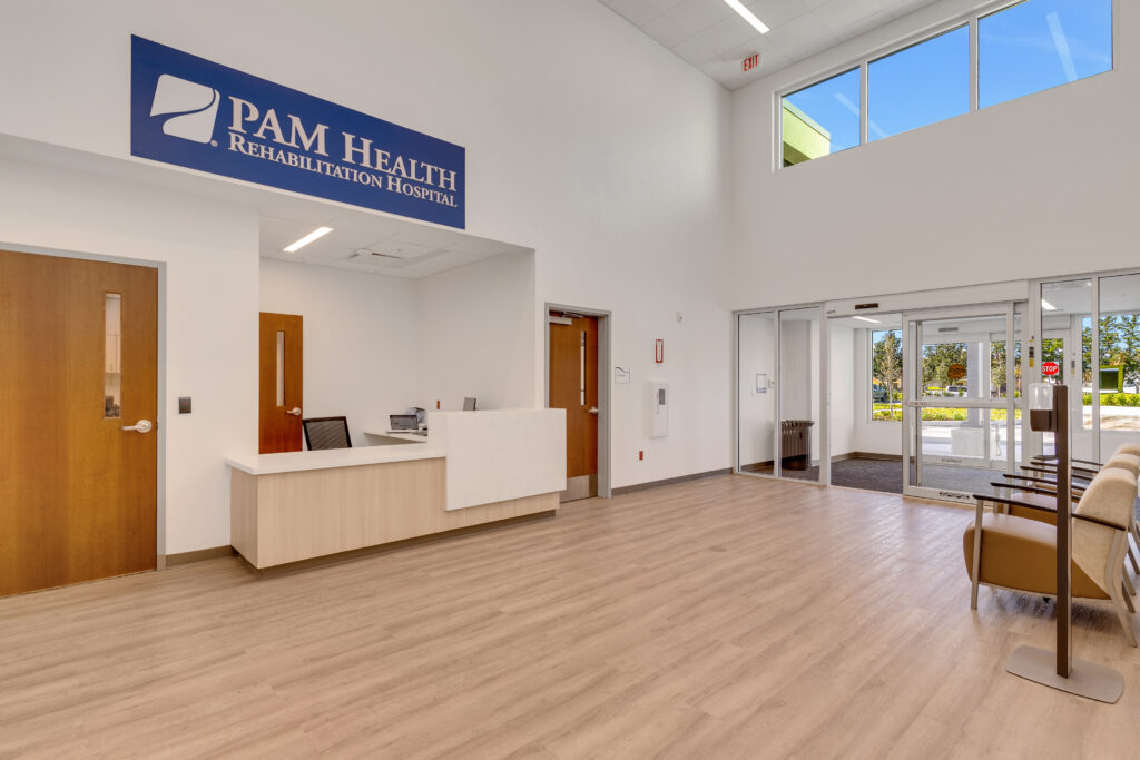 Reception area of PAM Health Rehabilitation Hospital with a check-in desk, wooden doors, and seating near a glass entrance.