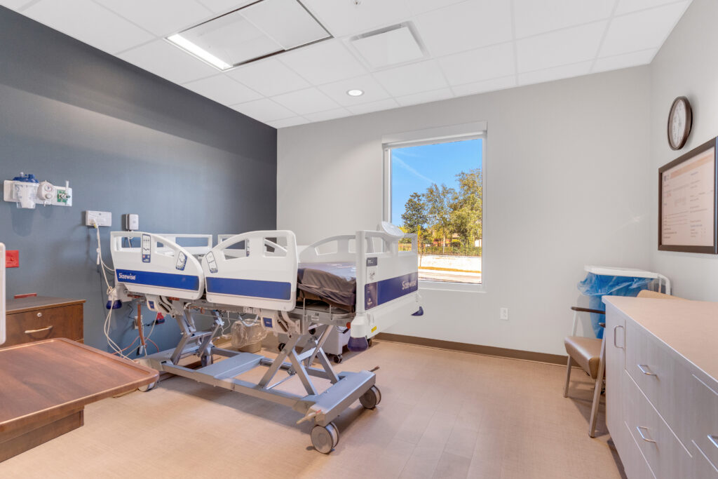 A hospital room featuring a central adjustable bed, medical equipment on the wall, a window overlooking trees, and a wooden cabinet.