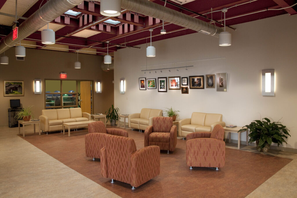 A waiting area featuring four patterned armchairs in the center, two beige sofas along the walls, potted plants, and framed artwork hanging on a white wall.