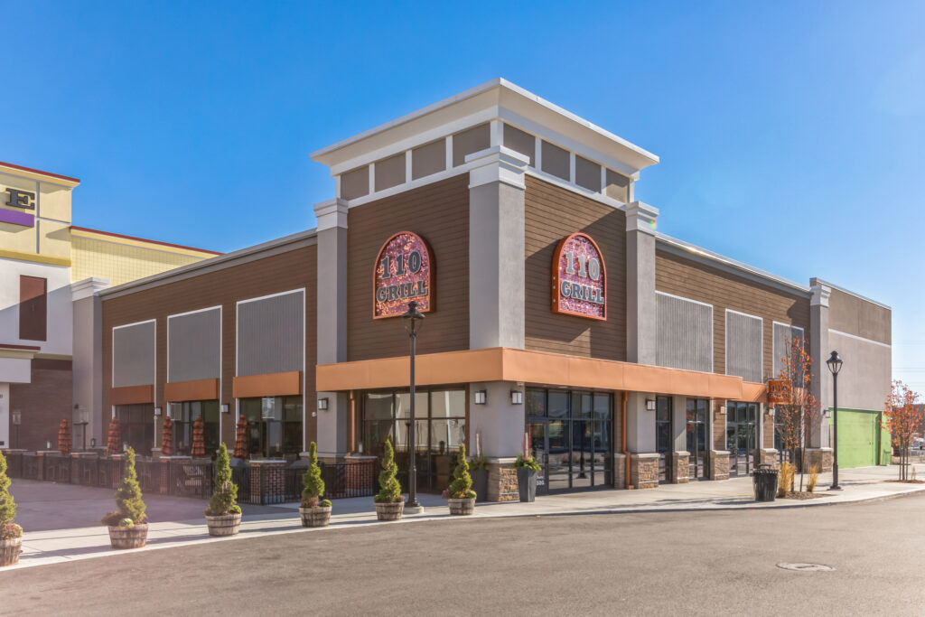 Exterior view of a 110 Grill restaurant with brown and grey siding, arched signage, and outdoor seating.