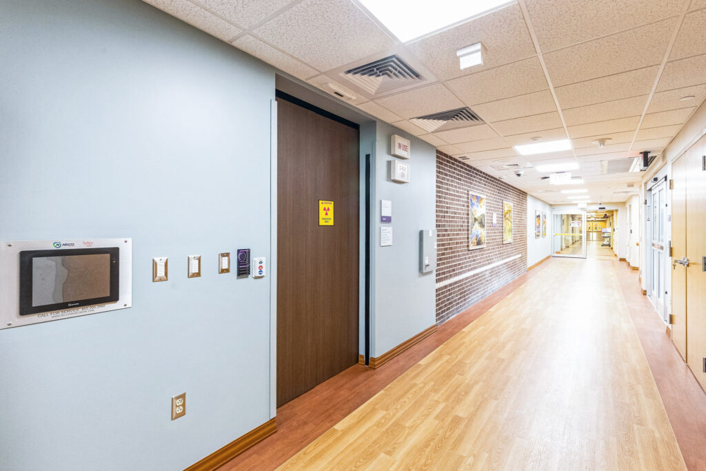A hospital corridor with wood flooring, a door marked with a radiation warning sign, and a touchscreen control panel on the wall.
