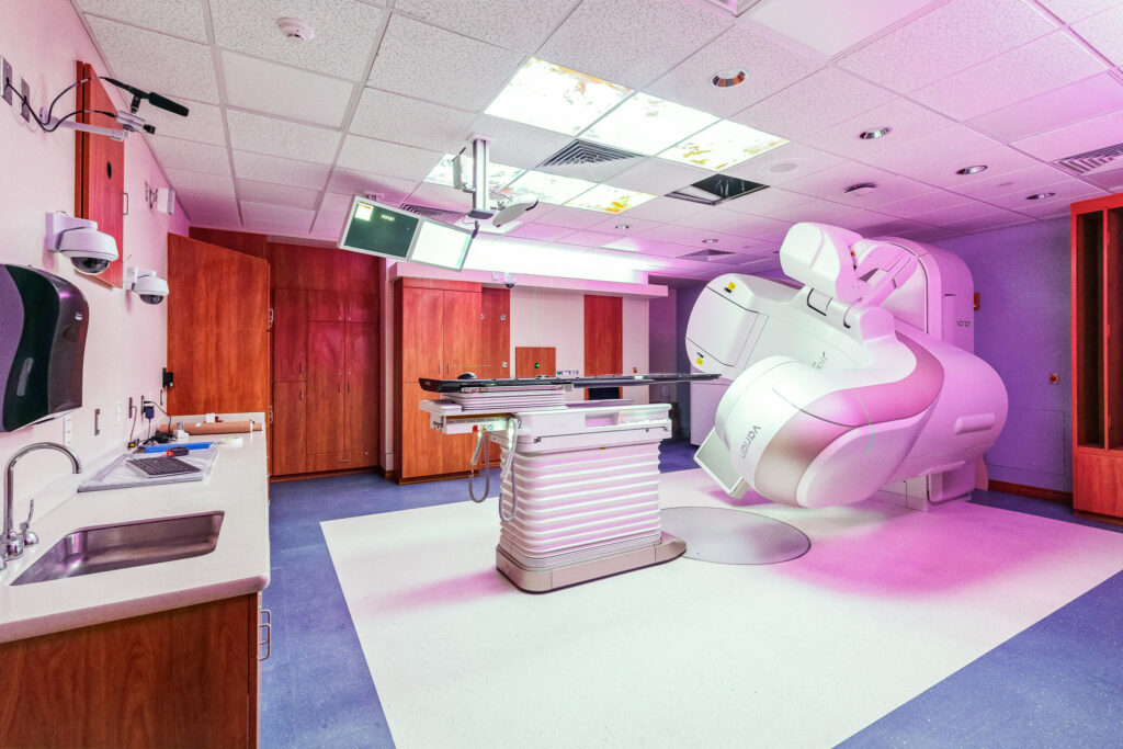 A medical treatment room featuring a Varian linear accelerator for radiation therapy positioned next to a patient table.