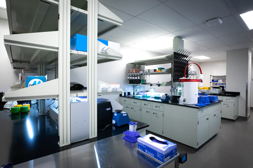 A laboratory interior featuring stainless steel work surfaces, cabinetry, and various scientific equipment including pipette racks, centrifuges, and test tube holders.