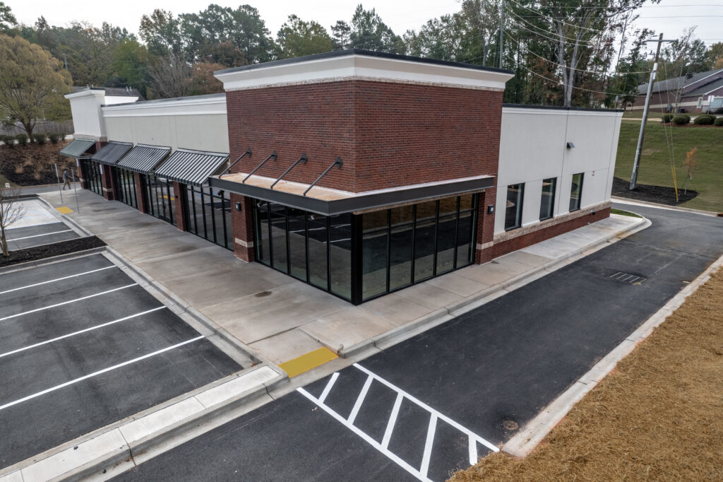 An aerial view of a vacant corner commercial building with large glass windows, a brick facade, and an adjacent parking lot.