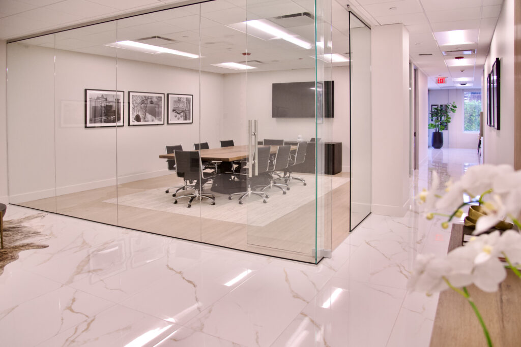 Glass-walled conference room featuring a long table with chairs, centered on a rug, with framed photos on the wall.