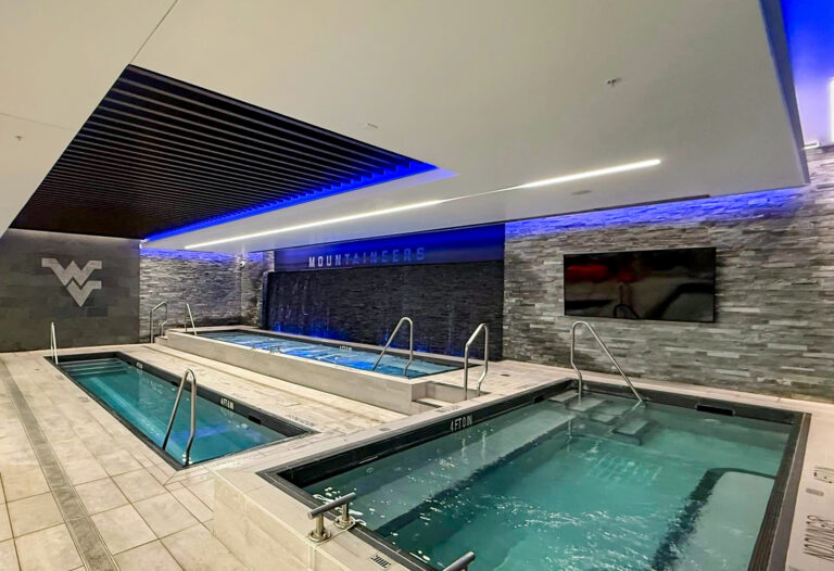 Three indoor hydrotherapy pools with metal handrails, blue accent lighting, and a West Virginia University logo on a stone wall.