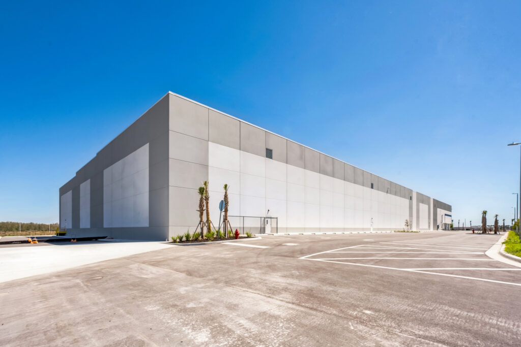 A large, grey warehouse building with a paved parking lot under a clear blue sky.