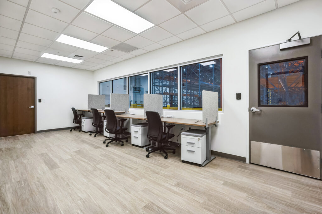An office workspace featuring three desks with rolling chairs and privacy dividers, facing a large window looking into a warehouse.