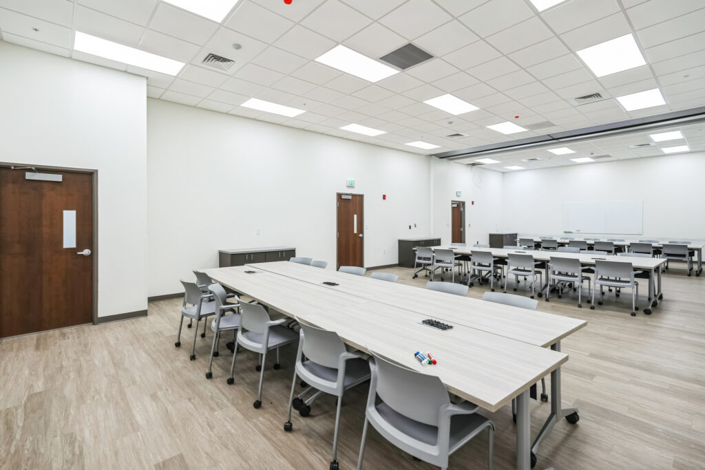 A bright, spacious conference room with rows of tables and grey chairs, multiple doorways, and a whiteboard.