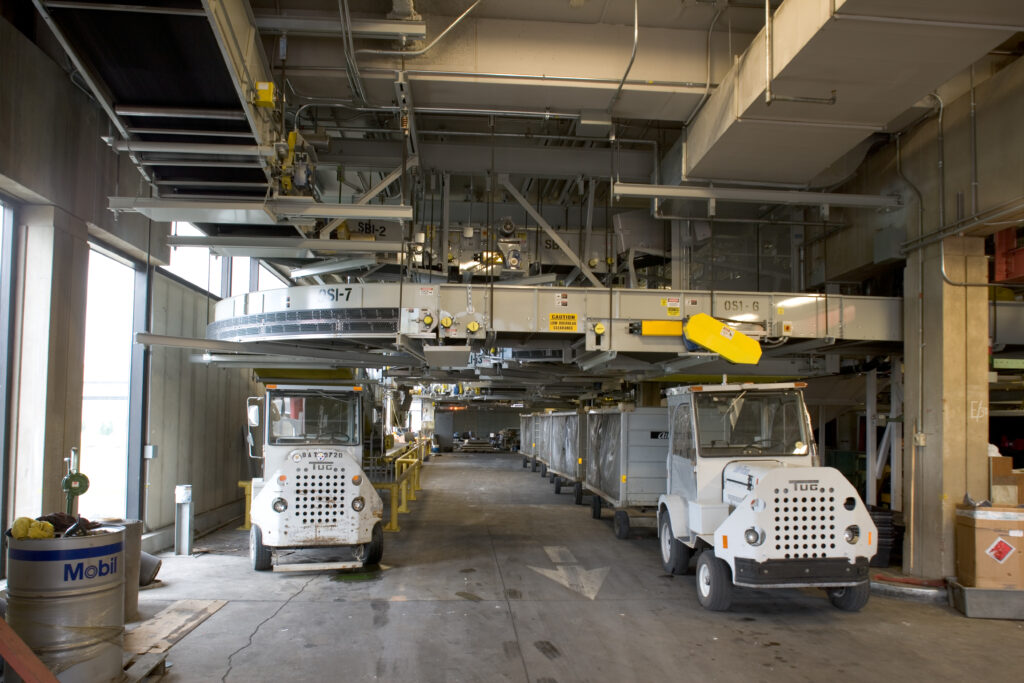 Two white airport baggage tugs parked under an overhead conveyor system in a maintenance bay.